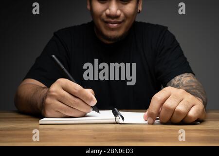 Asian Mann Schreiben auf einem Notizbuch mit Lächeln Gesicht auf Holztisch. Konzept über Remote-Arbeit. Stockfoto