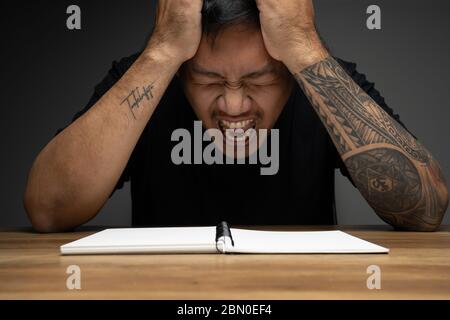 Asian Mann Schreiben auf einem Notizbuch mit wütenden Gesicht auf Holztisch. Konzept über Remote-Arbeit. Stockfoto