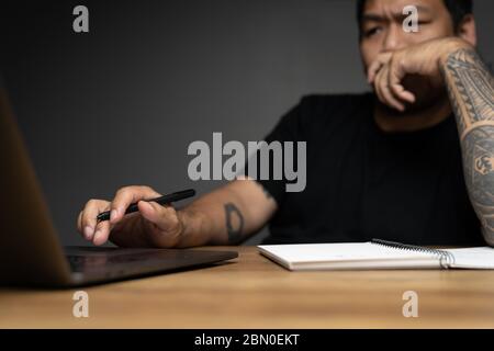 Asian Mann Blick auf Monitor mit Fokussierung Gesicht auf Holztisch mit Laptop. Konzept über Remote-Arbeit. Konzentrieren Sie sich auf die Hand. Stockfoto