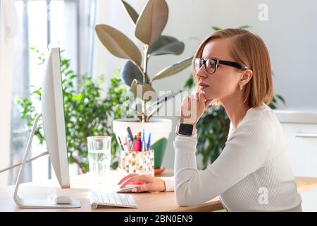 Nachdenkliche freiberufliche Frau in Brille, die am Computer von zu Hause aus während der Quarantäne durch Coronavirus arbeitet. Gemütlicher Arbeitsplatz umgeben von Pflanzen. Ein Stockfoto