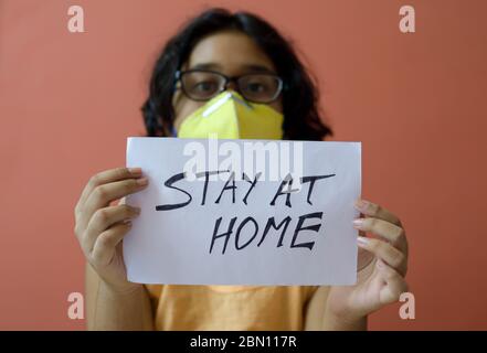 Kleines indisches Mädchen mit Gesichtsmaske hält ein Plakat in den Händen und zeigt eine Botschaft "Stay at Home" während der COVID-19 Pandemie, um soziale Distanz zu erhalten. Stockfoto