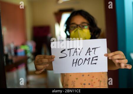 Kleines indisches Mädchen mit Gesichtsmaske hält ein Plakat in den Händen und zeigt eine Botschaft "Stay at Home" während der COVID-19 Pandemie, um soziale Distanz zu erhalten. Stockfoto