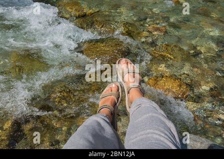 Gebräunte Beine der Frauen in offenen Sport-Sandalen auf einer Wanderung auf einem Bergfluss. Reisekonzept, das Konzept des Wanderens Stockfoto