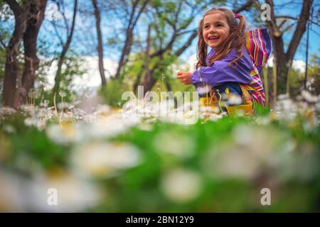 Glückliches kleines Mädchen, das nach dem Regen im Gras mit Blumen spielt. Sie trägt Regenmantel und gelbe Stiefel. Stockfoto