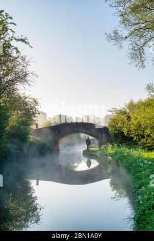 Mann, der an einem Frühlingsmorgen kurz nach Sonnenaufgang unter einer Ziegelbrücke am Oxford Kanal entlang des Wegs läuft. Upper Heyford, Oxfordshire, England Stockfoto