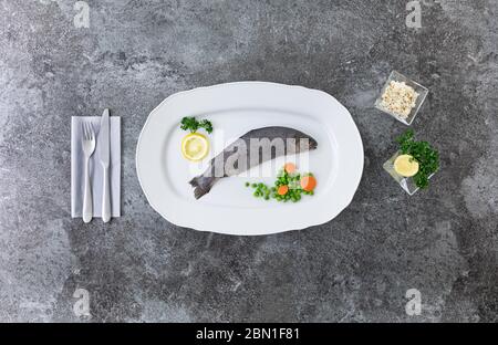 Eine ganze Regenbogenforelle auf einem weißen Servierteller mit Messer und Gabel, Zitronen, Petersilie, Erbsen und Karotten auf einem grauen Marmortisch Stockfoto