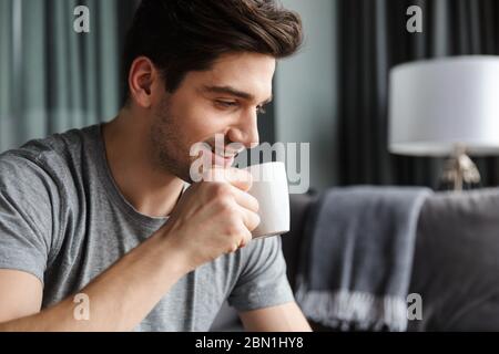 Nahaufnahme eines lächelnden jungen bärtigen Mannes, der Tee aus einer Tasse im Wohnzimmer trinkt Stockfoto