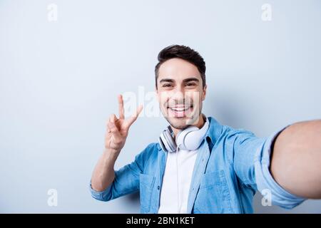 Selfie-Manie! Gutaussehend lächelnd junge Brunete Mann mit Stoppeln macht Selfie vor der Kamera und Gesturing Frieden auf hellblauem Hintergrund, in Jeans-Shirt Stockfoto