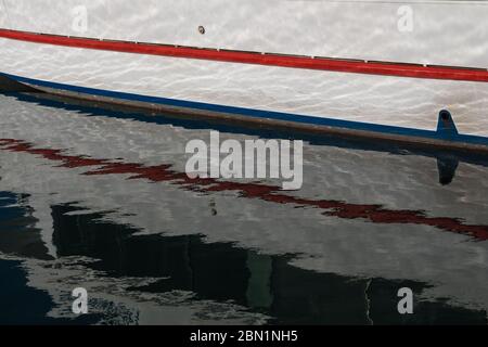 Weißes Boot mit roten und blauen Details in einem Hafen vertäut. Reflexion im Wasser, modifiziert durch kleine Wellen. Alghero, Sardinien, Italien. Stockfoto