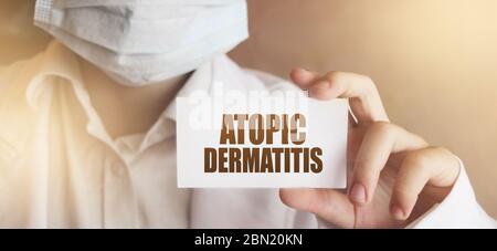 Atopische Dermatitis auf einer Karte, die von der Hand des Arztes gehalten wird. Arzt trägt chirurgische Medizin Gesichtsmaske. Medizinisches Konzept Stockfoto