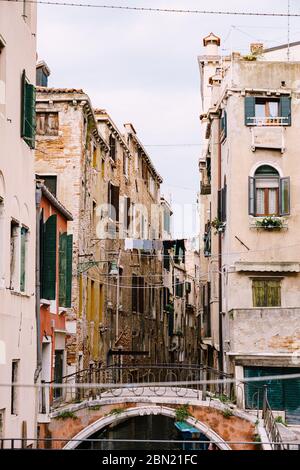 Nahaufnahmen von Fassaden in Venedig, Italien. Ein schmaler venezianischer Kanal, zwischen zwei Häusern, die Kleidung an einem Seil trocknen. Eine kleine Steinbrücke mit Metall Stockfoto