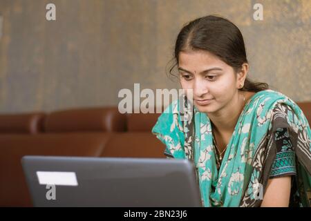 Lächelnde indische Mädchen Student oder Mitarbeiter beschäftigt auf Laptop sitzen zu Hause in lässigen Kleid, glückliche Frau studieren, E-Learning, mit Online-Software oder Stockfoto