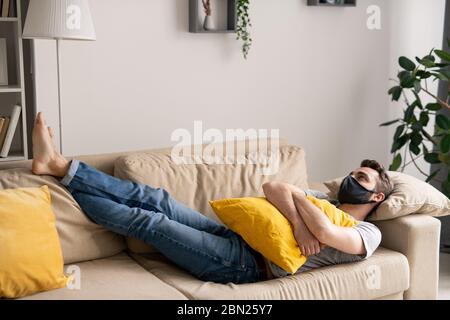 Junger Mann in Maske auf dem Sofa liegend und Blick auf die Decke, während aus Hause Isolation leiden Stockfoto