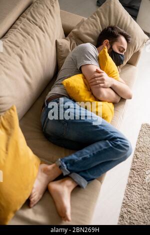 Oben Ansicht des verzweifelten jungen Mannes in schwarzer Gesichtsmaske mit Kissen auf Sofa in Selbstisolation schlafen Stockfoto