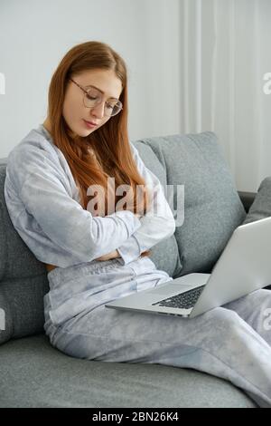 Ein rothaariges Mädchen in Brille und Pyjama sitzt mit einem Laptop auf der Couch, die Arme gefaltet und die Augen geschlossen. Stockfoto