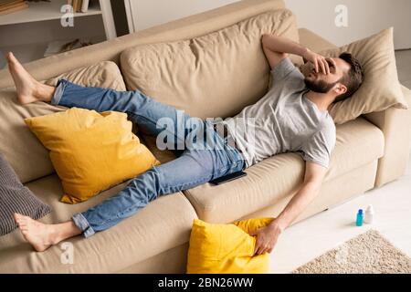 Oben Ansicht von traurig verwirrt jungen bärtigen Mann in Freizeitkleidung auf Sofa im Wohnzimmer liegen und Gesicht mit der Hand in Quarantäne Stockfoto