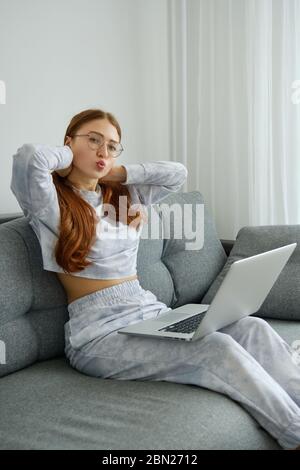 Ein rothaariges Mädchen in Pyjama und Brille sitzt halb auf der Couch mit einem Laptop auf ihrem Schoß und sendet einen Kuss. Stockfoto