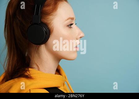 Kopfschuss. Ein rothaariges Mädchen mit Kopfhörern und einem gelben Hoodie steht im Profil auf blauem Hintergrund Stockfoto