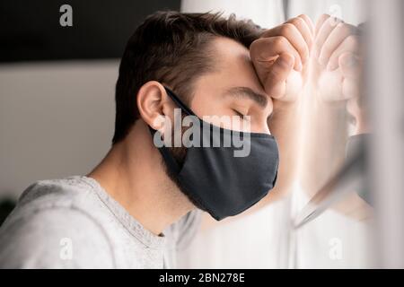 Junger Mann in Stoffmaske müde von der Isolation stehend mit geschlossenen Augen am Fenster, Quarantäne zu Hause Stockfoto