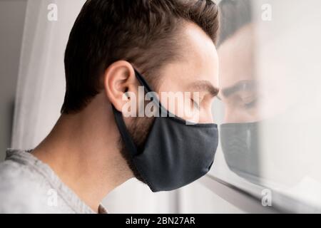 Traurig verwirrt junger Mann in Stoffmaske lehnte den Kopf auf Fenster und die Augen geschlossen, während Gefühl Depression wegen der Coronavirus Isolation Stockfoto