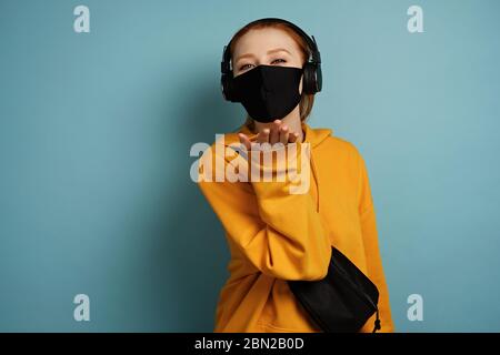 Ein Mädchen in einem gelben Hoodie und einer schwarzen Schutzmaske steht auf blauem Hintergrund und sendet einen Luftkuss. Stockfoto