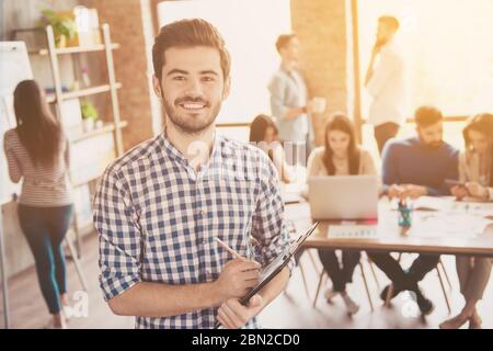 Alles für einen und einer für alle! Führung. Erfolgreiche stilvolle junge bärtige Mann in lässig karierten Hemd steht im Büro, lächelnd, machen Stockfoto