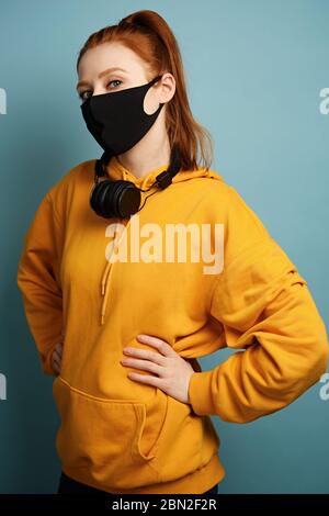 Rotschopf-Mädchen in einem gelben Hoodie und einer schwarzen Schutzmaske steht halb gedreht, hält die Hände an der Taille, schaut in den Rahmen Stockfoto