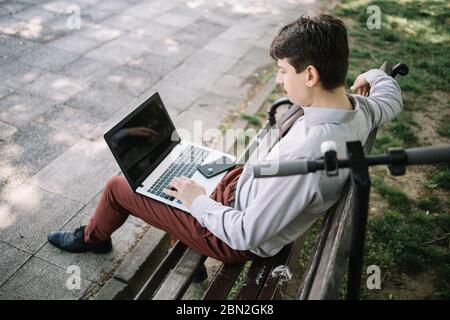 Seitenansicht des Mannes, der auf der Bank mit Laptop auf seinem Schoß ruht. Rückansicht des Mannes im Hemd, der auf Laptop arbeitet, während er auf Bank in der Nähe des Rollers sitzt. Stockfoto