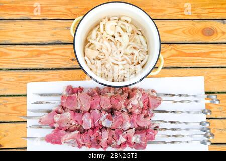 Flaches Lay. Frische rote Fleischstücke, die auf Metallspieße aufgereiht sind, liegen auf einem Tisch aus gelben Brettern. Das Fleisch wird mit Gewürzen eingelegt und zum Grillen vorbereitet. Stockfoto