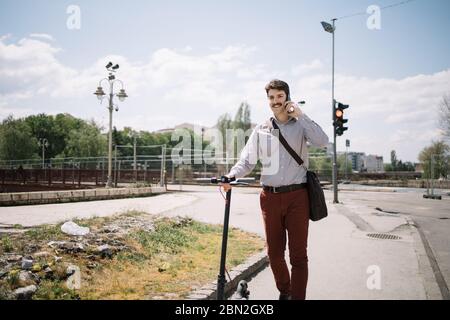 Glücklicher Geschäftsmann hält Roller und zu Fuß auf dem Bürgersteig. Lächelnder Mann, der auf der Straße läuft, während er den Roller hält und am Telefon spricht. Stockfoto