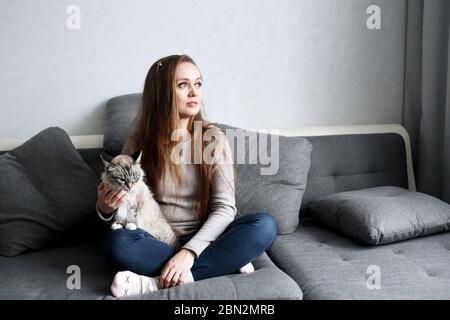 Junge trendige Frau sitzt auf der Couch zu Hause mit Hauskatze. Soziale Distanz, Isolation zu Hause Konzept. Ganzkörperportrait. Stockfoto