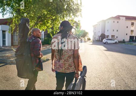 Männliche Musiker mit Gitarrenkoffer, die auf der sonnigen Straße spazieren Stockfoto