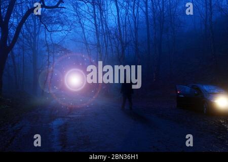 Ein Mann neben einem Auto, der auf ein glühendes UFO schaut, über einer Strecke in einem gruseligen nebligen Wald schwebt, Science-Fiction-Konzept. Stockfoto