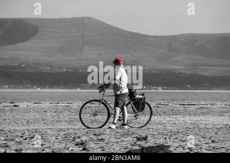 Mann in roter Mütze, britischer Urlauber, isoliert am leeren britischen Strand, der sein Fahrrad auf Sand schiebt. Monochromer Hintergrund. Urlaub im Sommer. Stockfoto