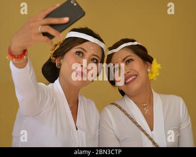 Zwei verkleidete, wunderschöne indonesische Balinesen in Weiß machen während einer religiösen Hindu-Tempelzeremonie ein Foto mit ihrem Handy. Stockfoto