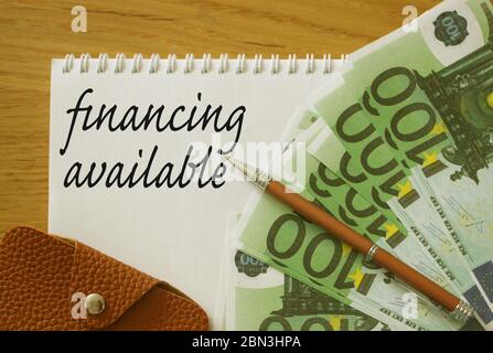 Finanzielle verfügbare Wörter auf Copybook-Seite, 100 Euro-Scheine, ein Stift und Leder-Brieftasche auf Holztisch. Sponsoring oder Versicherungszahlung für Unternehmen Stockfoto