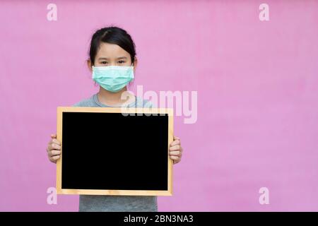 Kleine asiatische Mädchen in einer schützenden medizinischen Maske hält Tafel isoliert auf rosa Hintergrund. Schützen Sie sich vor Coronavirus oder Covid-19-Epidemie. Stockfoto