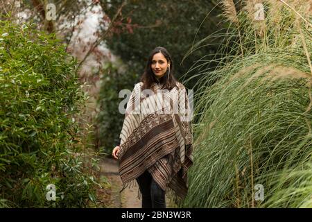 Junge Frau mit Poncho, die im Herbst zwischen hohen Gräsern geht. Stockfoto