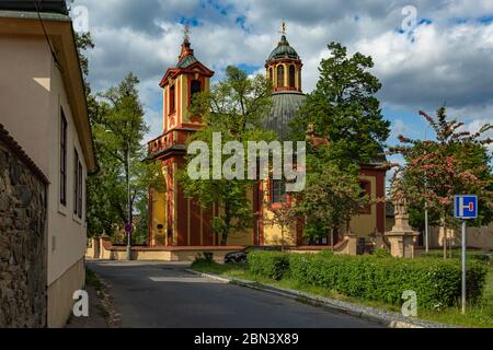 Kunratice, Prag / Tschechische Republik - 6. Mai 2020: Die barocke Kirche des Hl. Jakobus des Großen mit rot-gelber Fassade, umgeben von grünen Bäumen. Stockfoto