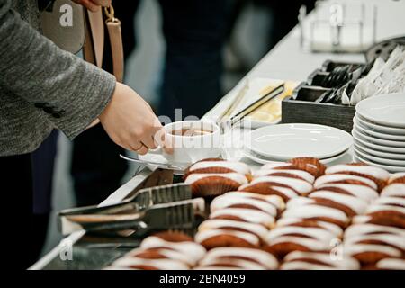 Eine Frau in einer grauen Jacke, deren Gesicht nicht zu sehen ist, nimmt eine Tasse Tee mit Zitrone von einem Banketttisch, auf dem gebrühten Kuchen und einem Stapel Weiß liegen Stockfoto