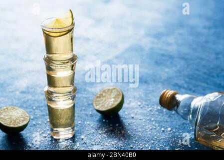 Goldener Tequila in Schnapsglas mit Salz und Limette, auf blauem Tisch, Hintergrundbeleuchtung. Drei Tequila-Schüsse gestapelt. Mexikanisches traditionelles Getränk. Stockfoto