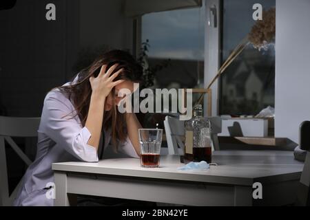 Depressive müde Ärztin sitzt am Tisch zu Hause hält die Hände hinter dem Kopf und trinkt starken Alkohol nach der Arbeit im Krankenhaus während Coronavirus Stockfoto