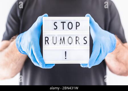 Fake News Infosemics during Covid-19 Pandemie Concept. Mann trägt Schutzmaske und medizinische Handschuhe an den Händen halten Leuchtkasten mit Text Stop Rumors. Stockfoto