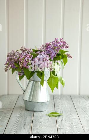 Metallgiesskanne mit einem Bouquet von Flieder auf einem hölzernen Hintergrund nahe dem Fenster. Stillleben im Frühling. Stockfoto