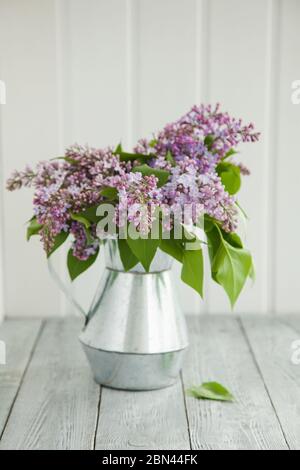 Metallgiesskanne mit einem Bouquet von Flieder auf einem hölzernen Hintergrund nahe dem Fenster. Stillleben im Frühling. Stockfoto