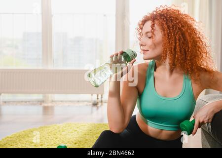 Schöne junge Frau Trinkwasser aus Sportflasche und halten grünen Hantel Gewicht während des Heimtrainings Training Training in covid Isolation Periode Stockfoto