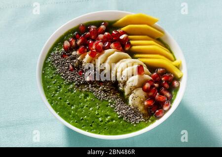 Schüssel mit grünem Smoothie mit geschnittenen Mango, Banane, Granatapfelkernen und Chiasamen, Ahornsirup für ein gesundes Frühstück am Morgen Stockfoto