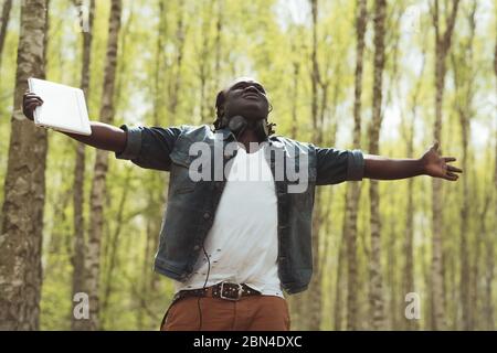 Afrikaner mit Notebook und Kopfhörer mit ausgestreckten Händen genießen die frische Luft im Wald Stockfoto