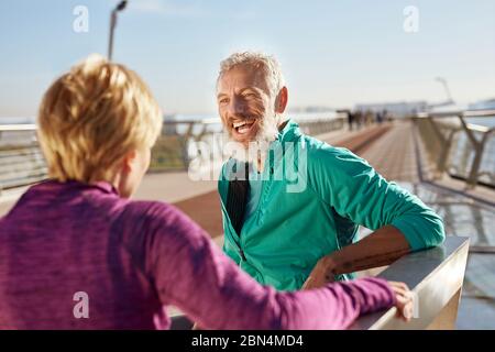 Nach dem Morgen laufen. Fröhlicher reifer bärtiger Mann in Sportkleidung, der mit seiner Frau spricht und am Morgen auf der Brücke lächelt Stockfoto