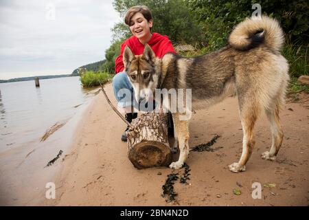 Kurzhaarige Mädchen in einem roten Hoodie und Jeans spielt mit einem Husky Hund am Flussufer Stockfoto